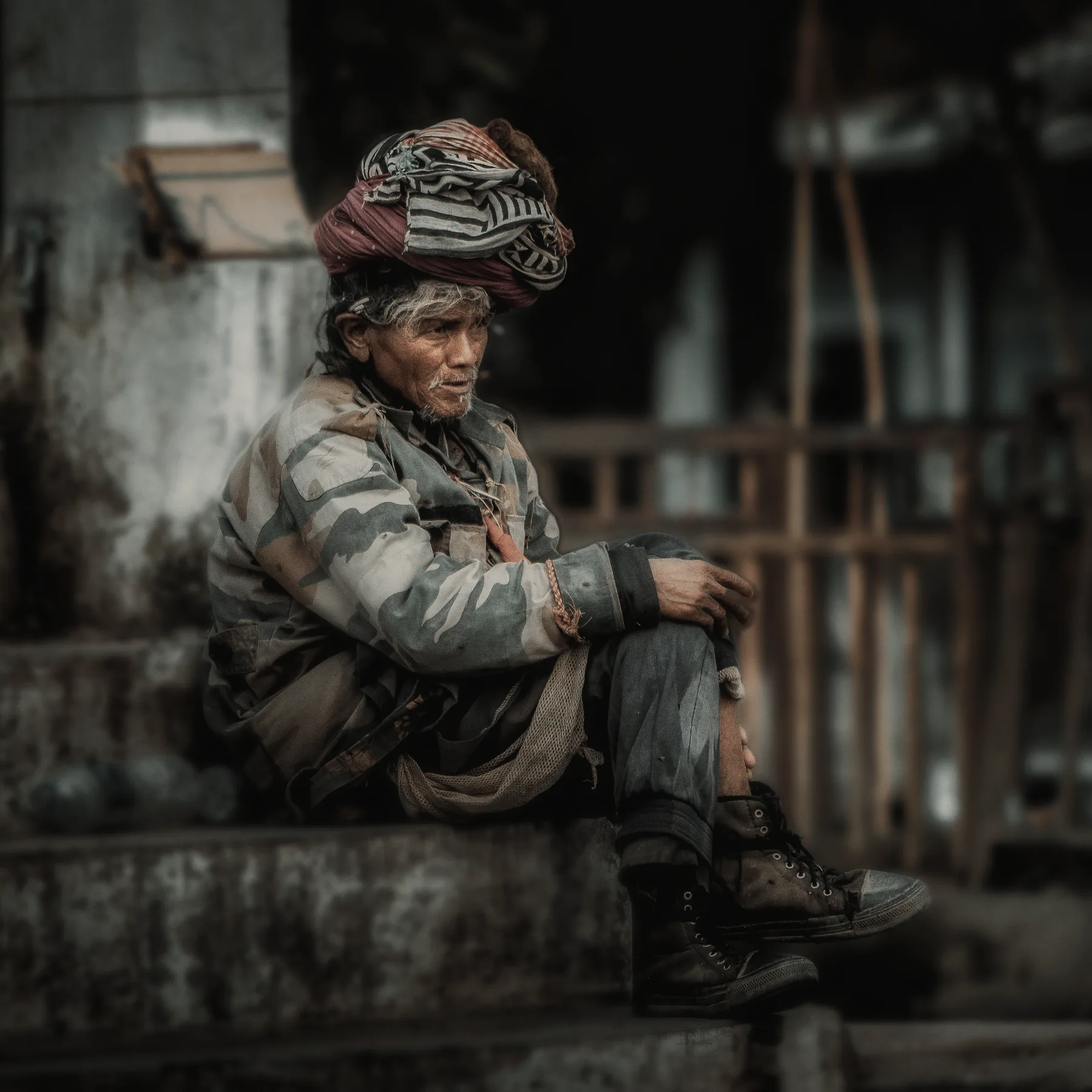 An elder sitting on a step in the market, wearing a patterned turban and camouflage coat